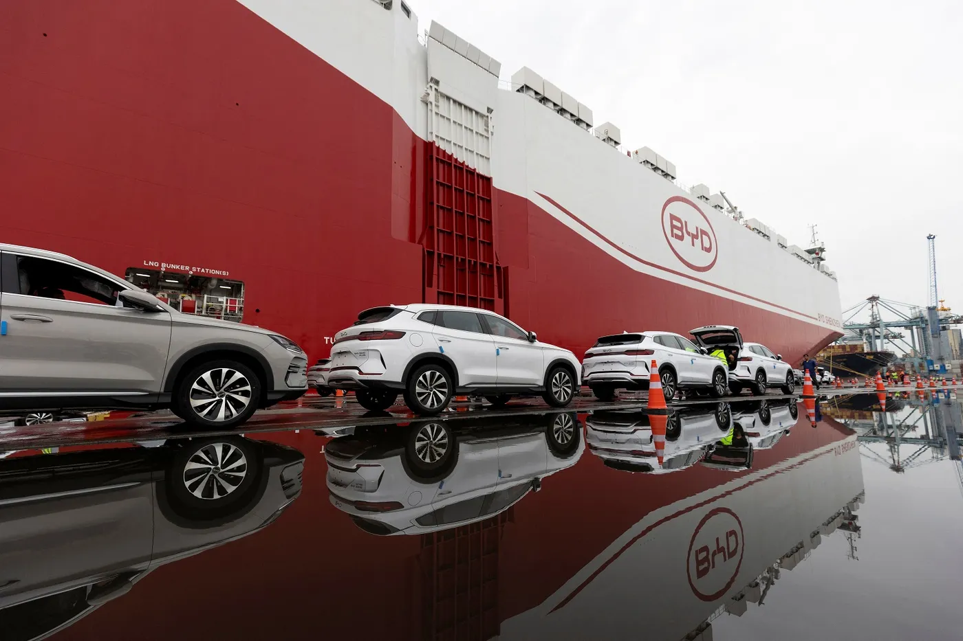 Cars lined up for loading onto a large BYD cargo ship, with reflections visible on the wet ground.
