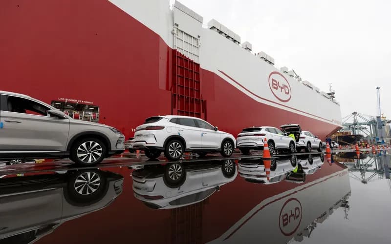 Cars lined up for loading onto a large BYD cargo ship, with reflections visible on the wet ground.