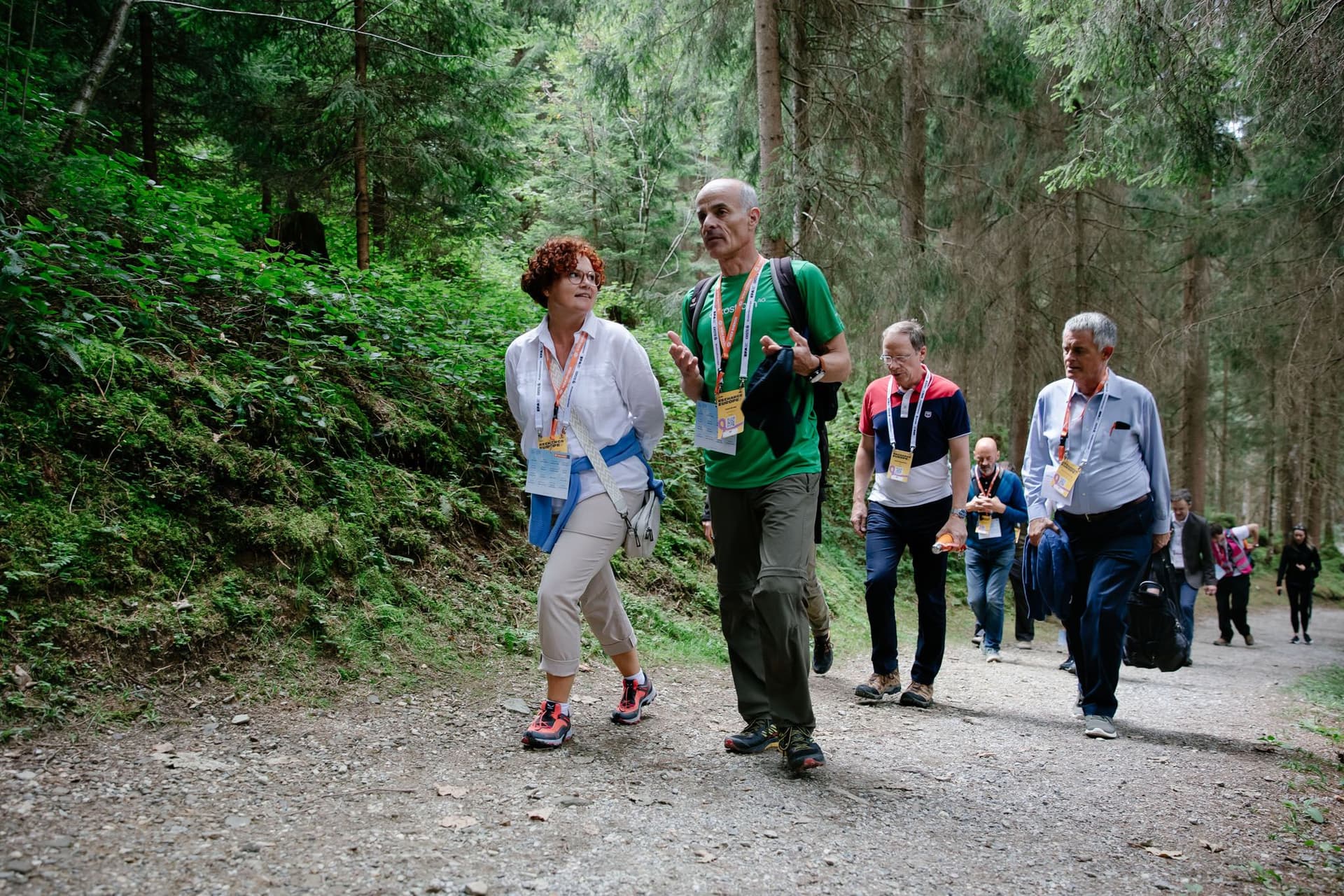 Eine Frau geht mit einem Mann durch den Wald, sie reden miteinander. Dahinter folgen mehrere Personen.