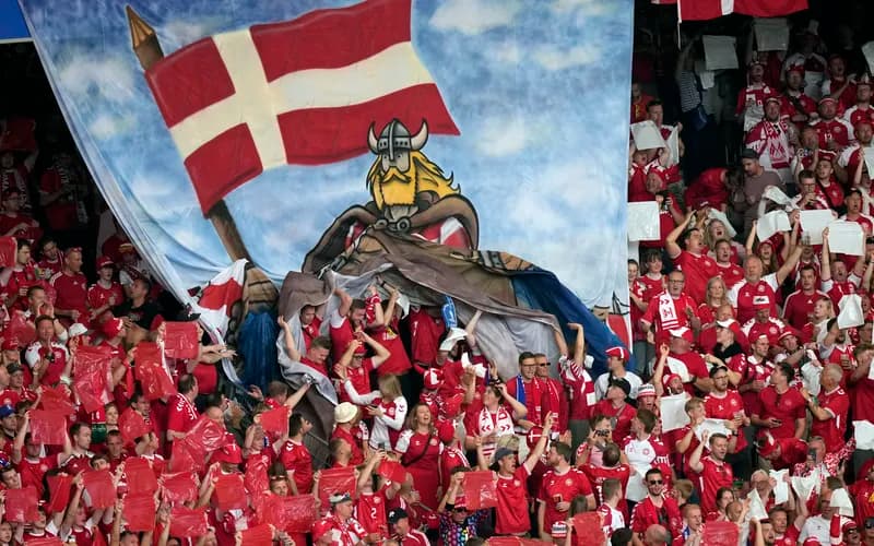 Fans in red shirts holding flags and banners beneath a large mural of a Viking with a Danish flag, creating a lively atmosphere in the stadium.