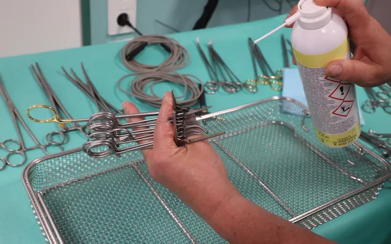 A person cleaning surgical instruments with disinfectant spray on a tray, surrounded by various medical tools on a teal surface.