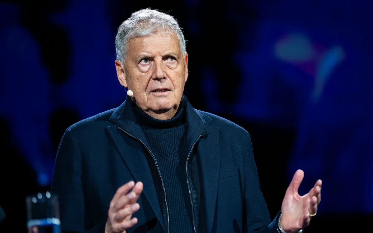 An older man with a microphone headset speaks passionately on stage, wearing a dark jacket against a dark blue background.