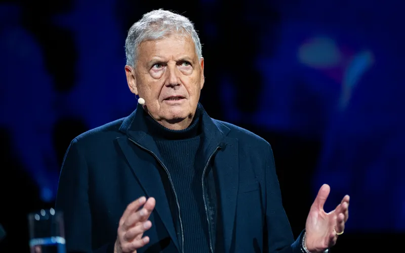 An older man with a microphone headset speaks passionately on stage, wearing a dark jacket against a dark blue background.