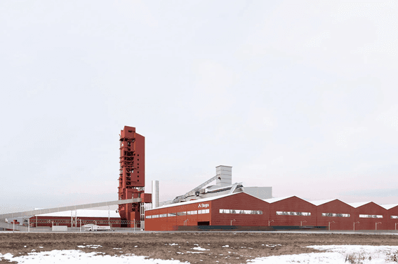 Industrial facility with red and white buildings, featuring a tall red tower, conveyor belts, and a snowy foreground under a cloudy sky.